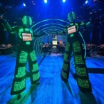 Two performers in green glowing LED robot stilt suits