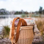Woven wicker picnic basket with shoulder strap and blanket sitting outdoors near a lake.