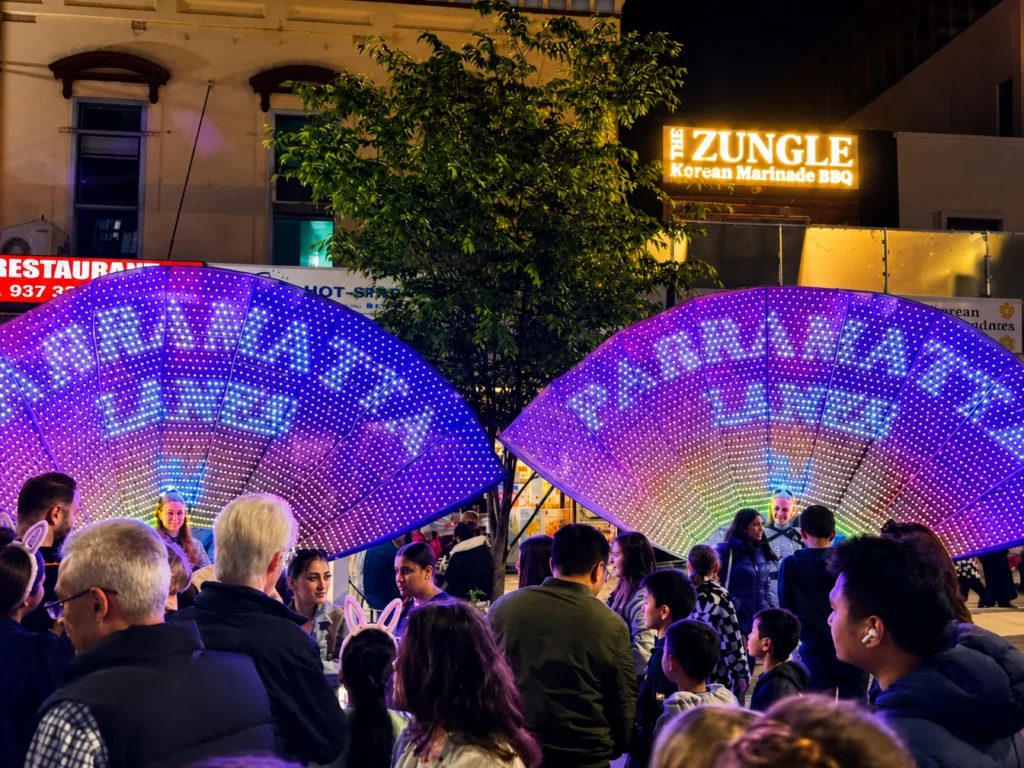 Large glowing LED fan structures displayed above outdoor street crowd
