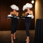 Two women wearing flower hats and black dresses serving drinks