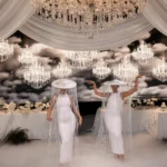 Dancers wearing white dresses and fringed tassel hats performing indoors.