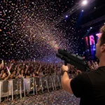 Person firing a confetti gun over a cheering concert crowd.