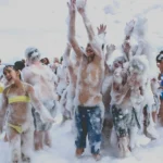 Excited crowd covered in white foam raising hands at a daytime event.