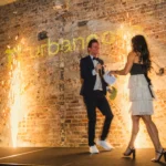 Man and woman walking on stage in front of cold spark pyrotechnics and brick wall.
