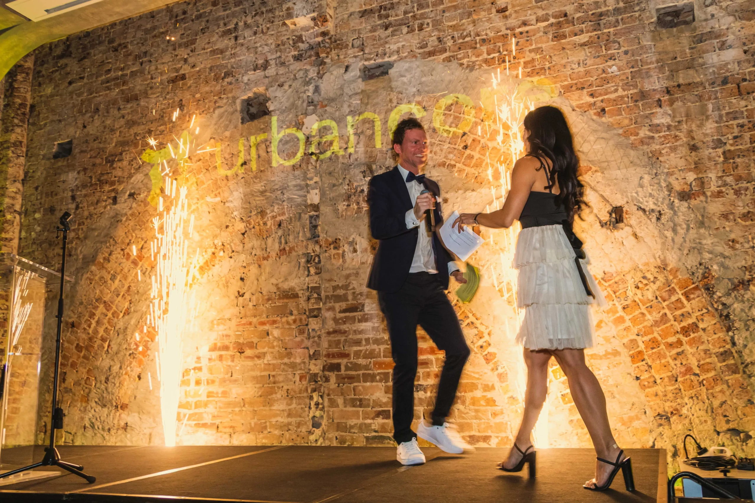 Man and woman walking on stage in front of cold spark pyrotechnics and brick wall.