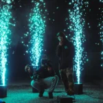 Two men on stage framed by vertical cyan cold spark fountains at night.