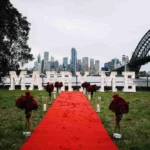 Red carpet and MARRY ME marquee letters set up on a pier with a city skyline.